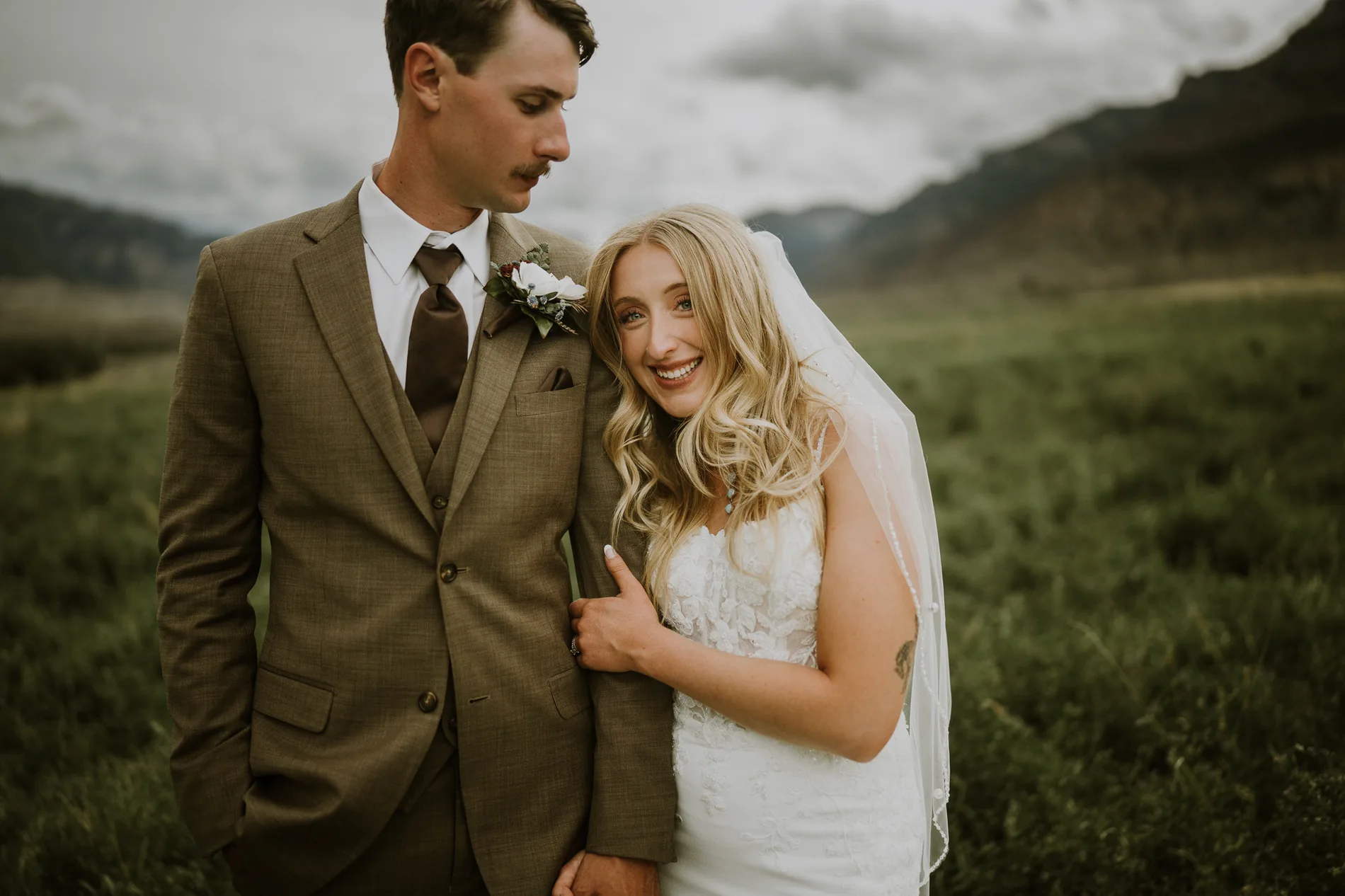 Blonde bride smiling at the camera alongside the groom in a brown suit